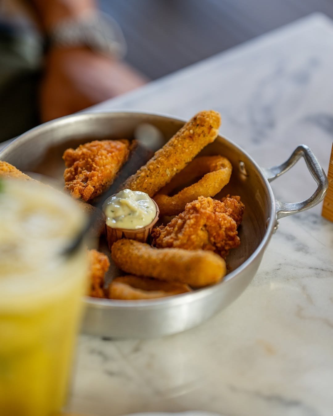 Taça de metal com sticks de mozzarella e outros fritos, com molho branco, ao lado de um copo de limonada amarela