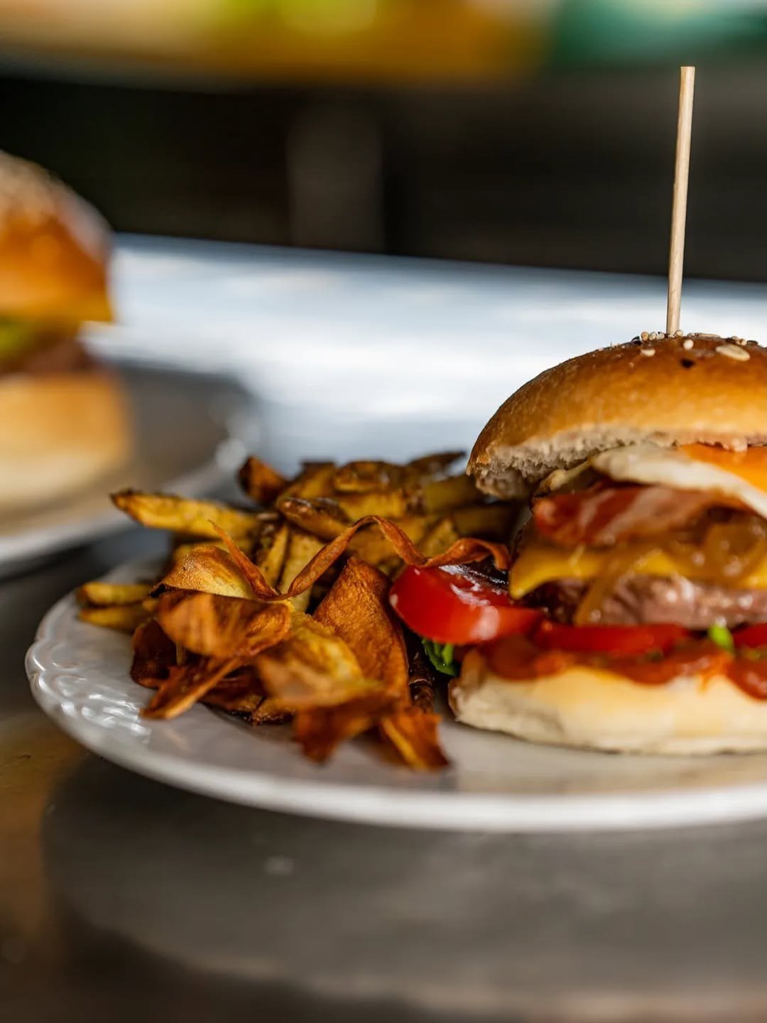 Hambúrguer com pão de sementes e batatas fritas num prato branco, com um segundo hambúrguer desfocado ao fundo
