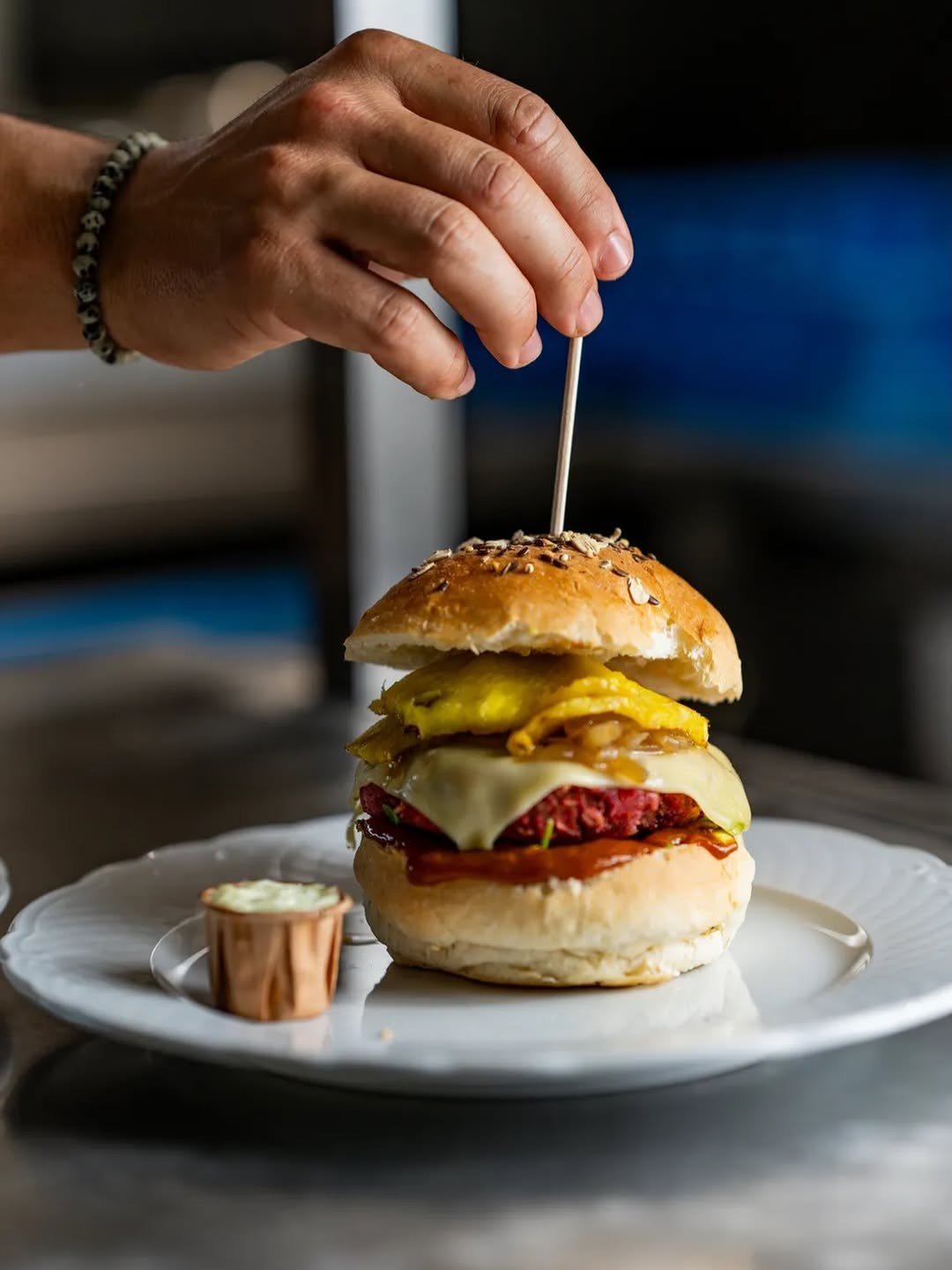 Mão a espetar um palito num hambúrguer alto com queijo cheddar e carne, servido num prato branco